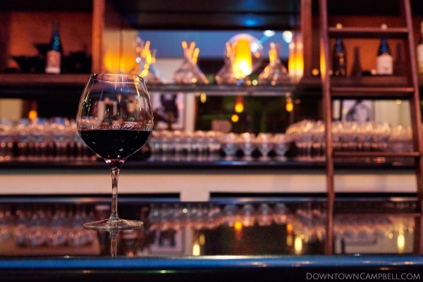 Large wine glass filled with red wine sitting alone on a wine bar with a wine library in the background.